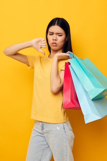 色とりどりの買い物袋が付いている黄色のTシャツのアジアの外観を持つ女性は、変更されていない背景を分離しました