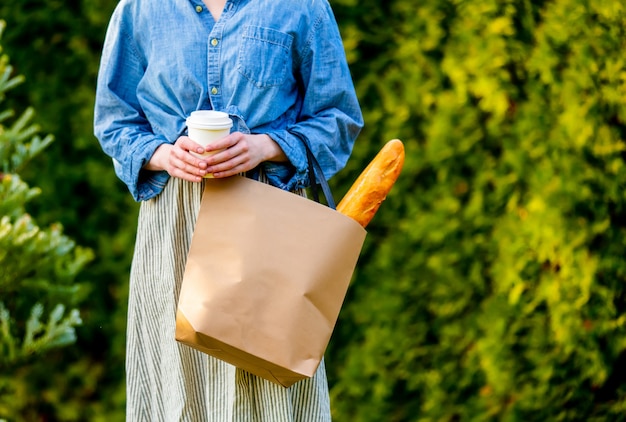 ショッピングバッグにバゲットと庭で一杯のコーヒーを持つ女性