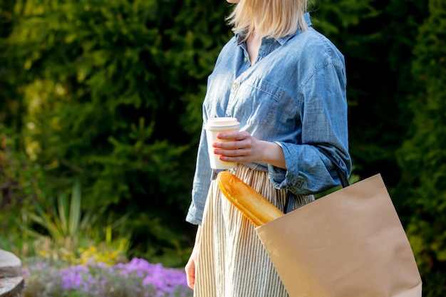 ショッピングバッグにバゲットと庭で一杯のコーヒーを持つ女性