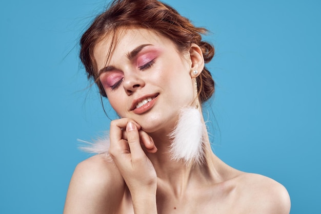 Woman with bare shoulders fluffy earrings bright makeup blue background