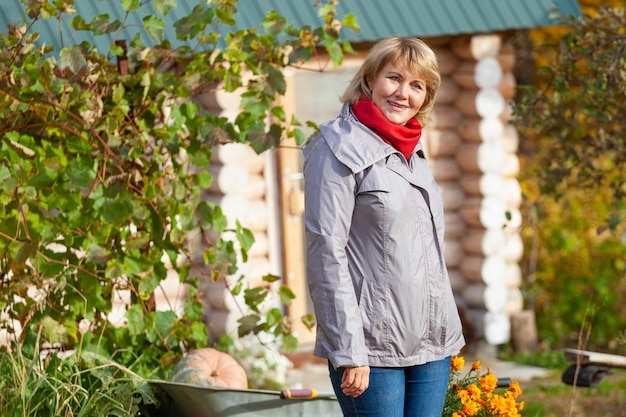 A woman with a basket is in the garden. A middle-aged woman in jeans and a jacket holds flowers against autumn trees. She's harvesting.