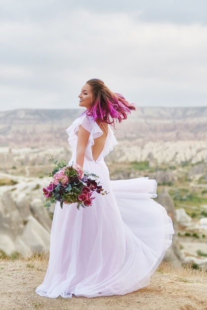 그녀의 손에있는 꽃의 아름 다운 부케와 여자 새벽 일몰의 광선에 산에서 춤을. 여자 몸에 아름다운 흰색 긴 드레스. 분홍색 머리 춤을 가진 완벽한 신부
