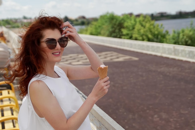 美しい赤い髪とサングラスで川沿いにアイスクリームを食べる女性