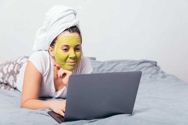 얼굴에 미용 마스크를 쓴 여자가 노트북을 들고 침대에 누워 있다