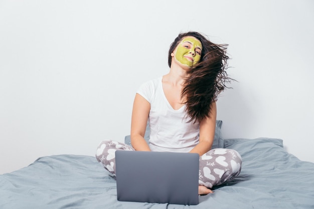 Женщина с маской красоты на лице сидит на кровати с мокрыми волосами