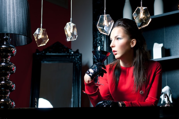 Woman with black hair in red dress and leather gloves sitting in restaurant and drinking cocktail from martini glass