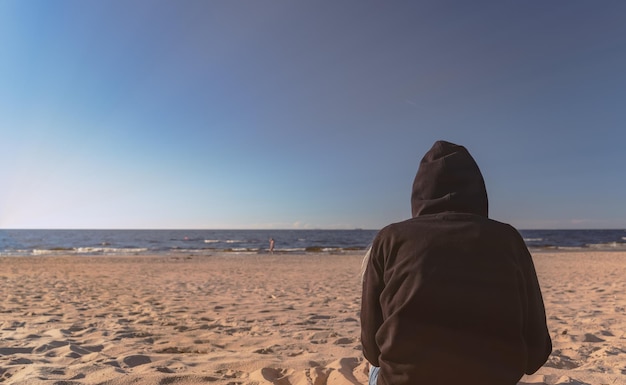 La donna con la felpa con cappuccio nera sull'essere umano della spiaggia si siede nella sabbia e guarda dentro