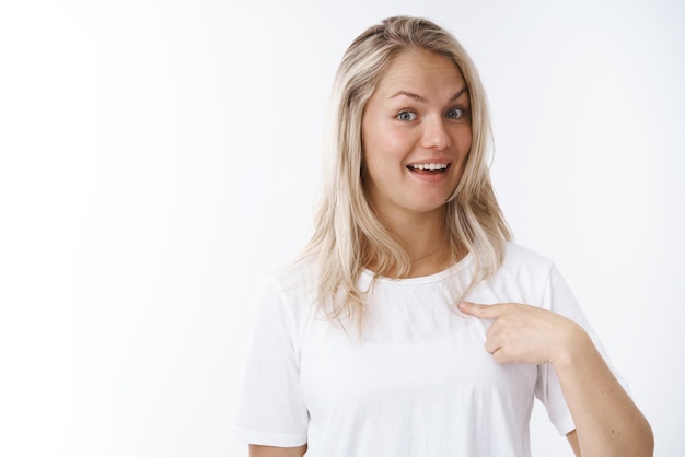 woman with blond hair in white t-shirt raising eyebrows in surprise smiling delighted pointing herself