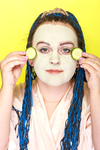 Woman with blue afro braids face in a frozen mask of green clay with pieces of cucumbers in her hands on a yellow surface