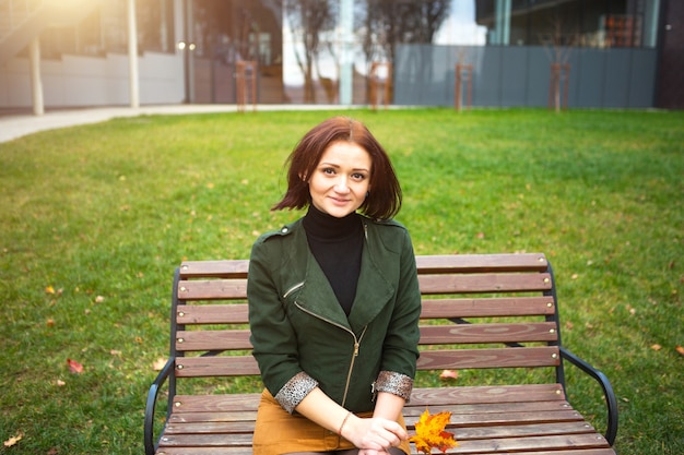 A woman with a bob haircut and a yellow fallen maple leaf in a green jacket and a mustard mini skirt. Autumn portrait on a park bench in a closed position, leg on leg. Autumn mood and fashion