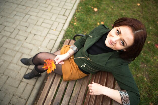 A woman with a bob haircut and a yellow fallen maple leaf in a\
green jacket and a mustard mini skirt. autumn portrait on a park\
bench in a closed position, leg on leg. autumn mood and\
fashion