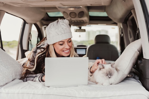 Woman with a bobble hat Using Laptop While Lying With cat In Camper Trailer Digital nomad lifestyle