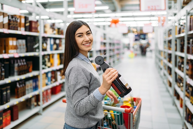 Donna con bottiglia di bevanda alcolica in supermercato, reparto bevande alcoliche, acquisti in famiglia. cliente femminile che sceglie vino in negozio, acquirenti nel mercato