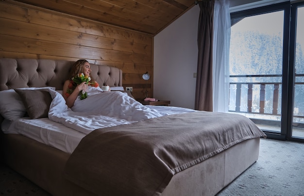 Женщина с букетом цветов, лежащая на кровати и наслаждающаяся праздничным утром с ароматной чашкой кофе во время отдыха в уютной спальне в гостиничном номере с красивым видом