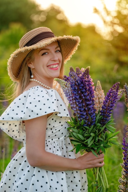 woman with  bouquet of lupines in polka dot dress and a straw hat walks on lawn with lupin at sunset