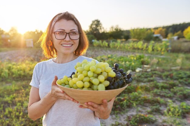 Фото Женщина с миской свежесобранного синего и зеленого винограда, солнечный фон сада. садоводство, сельское хозяйство, виноградник, сбор урожая, концепция здоровой натуральной органической пищи