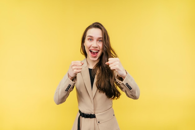 woman with braces on her teeth and in a jacket rejoices with clasped hands on a yellow background