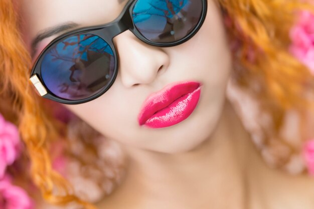 A woman with bright orange hair and sunglasses with a blue rimmed eyeglasses