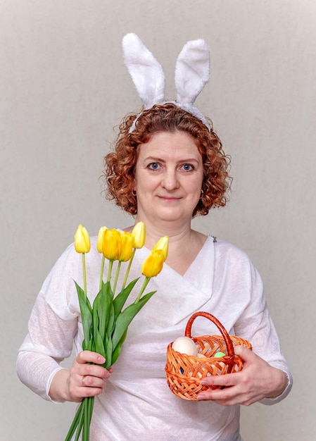 A woman with bunny ears holds a basket with Easter eggs and a bouquet of yellow tulips