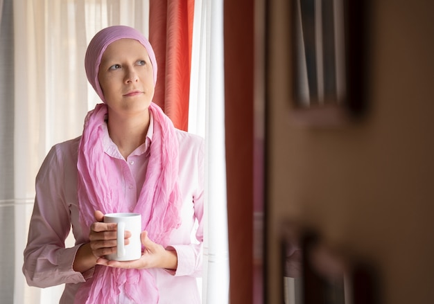 Woman with cancer and pink scarf on her head drinking coffee and looking out the window