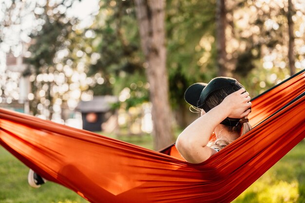 Woman with cap resting in comfortable hammock during sunset Relaxing on orange hammock between two trees pine enjoying the view