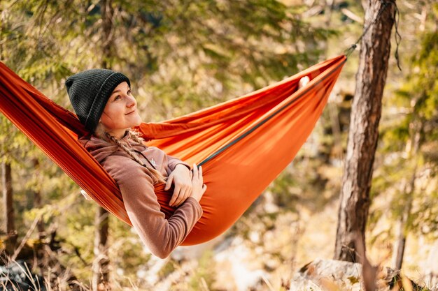 Woman with cap resting in comfortable hammock during sunset Relaxing on orange hammock between two trees pine enjoying the view