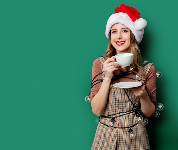 Woman with christmas lights and cup of coffee on green\
wall