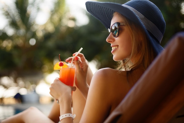야외용 데크 의자에 있는 수영장 근처의 열대 태양 아래서 칵테일 잔을 들고 있는 여성