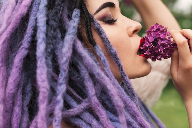 woman with color dreadlocks is touching lilac flowers with her lips