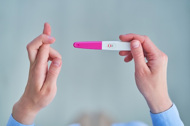 Woman with crossed fingers holds a positive pregnancy test with two stripes