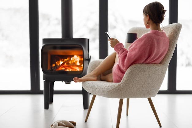 自然の家の暖炉のそばの椅子にカップを持つ女性