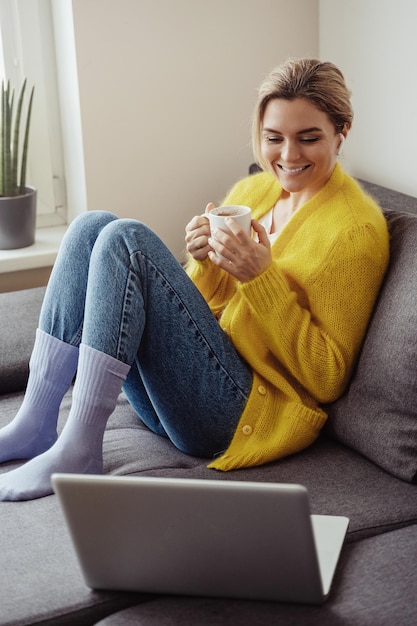 Woman with cup of hot coffee sitting on the sofa and using\
laptop computer at home