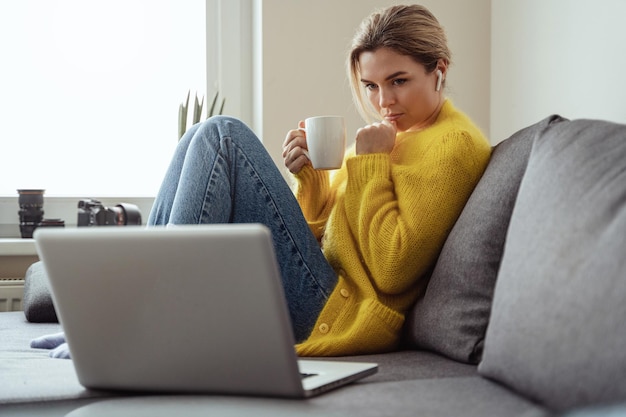 Woman with cup of hot coffee sitting on the sofa and using laptop computer at home