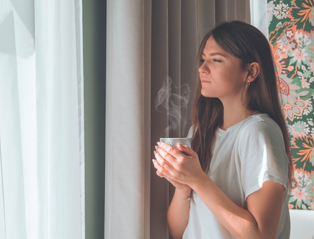 窓際に温かい飲み物のカップを持つ女性。窓を見てお茶を飲む。おはようお茶で。秋冬時間