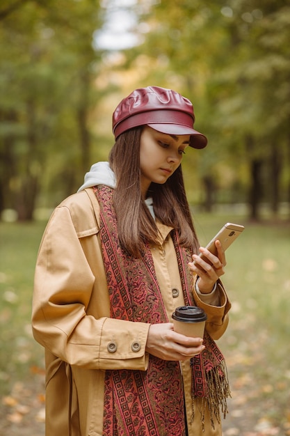 秋の公園でスマートフォンを使って持ち帰り用のコーヒーを飲む女性