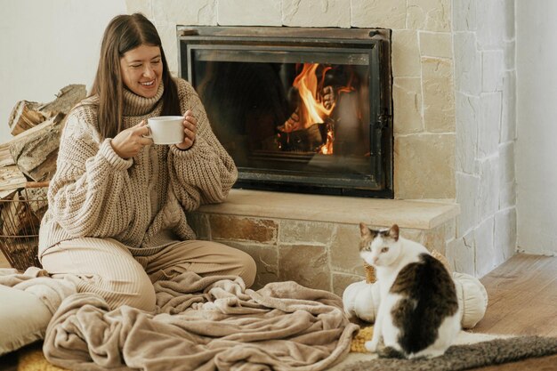 Photo woman with cup of warm tea and cat relaxing at fireplace autumn hygge heating house