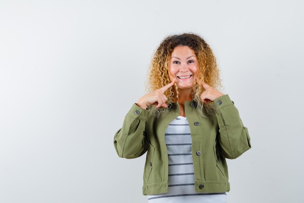 Photo woman with curly blonde hair pointing at her smile in green jacket and looking cheerful. front view.