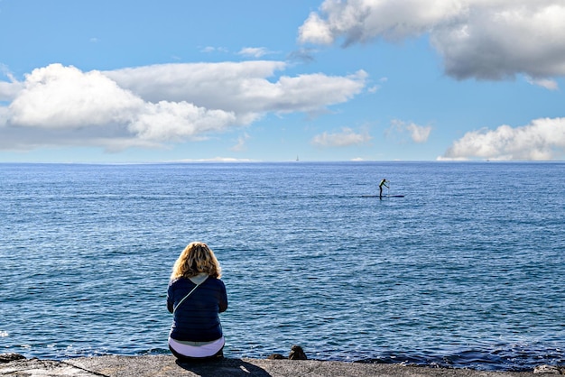 Woman with curly blonde hair, sitting on the beach jetty looking at the horizon of the sea.