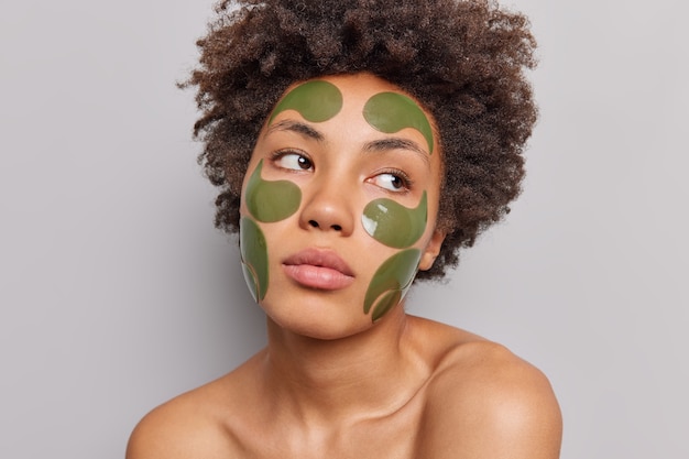 Woman with curly hair applies green beauty patches stands bare shoulders on grey