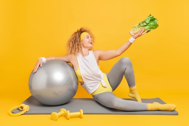 woman with curly hair dressed in activewear holds green vegetable poses with fitness ball dumbbells and headphones 