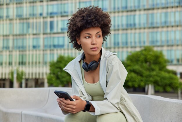 woman with curly hair dressed in sportswear messages in social networks browses content publication during break after training poses in urban setting