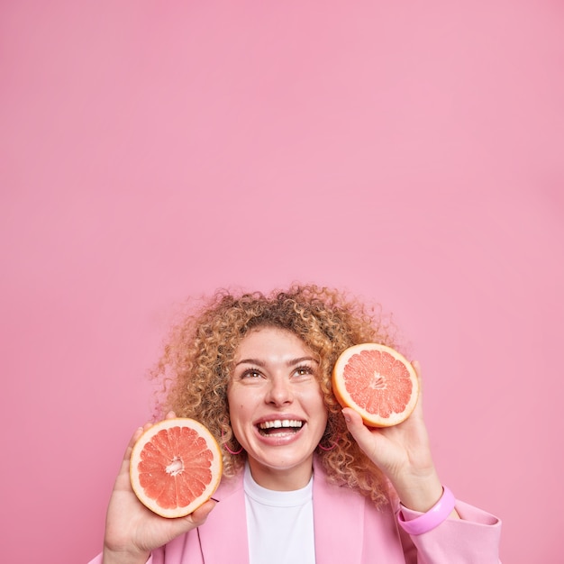 woman with curly hair holds fresh grapefruit slices smiles broadly dressed formally concentrated overhead 