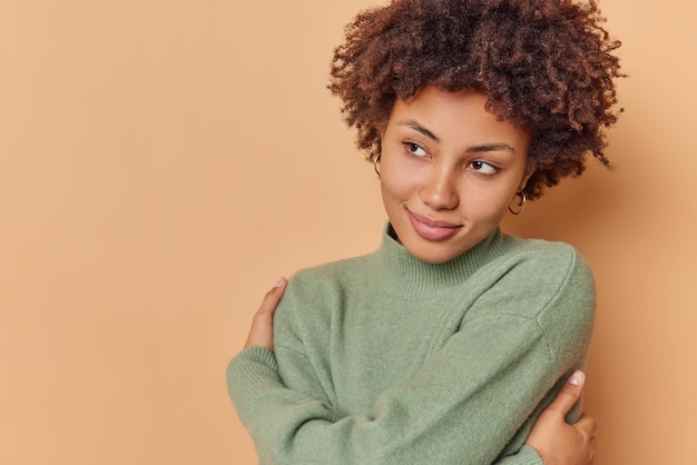 woman with curly hair hugs herself enjoys soft warm jumper looks thoughtfully away poses delighted against beige blank copy space for your advertising content