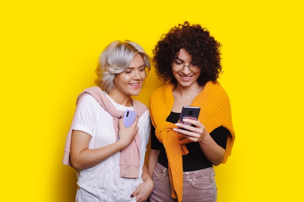 Woman with curly hair looks at something on the phone while woman with blond and short hair peeks ye...