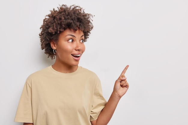 La donna con i capelli ricci sembra sorprendentemente lontana tiene la bocca aperta indica nell'angolo in alto a destra indossa una maglietta casual isolata su un muro bianco