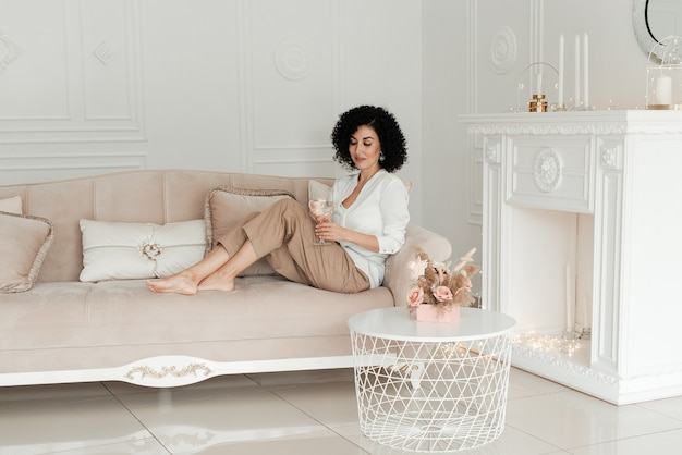 Woman with curly hair sits on the sofa with legs and holds a glass of water