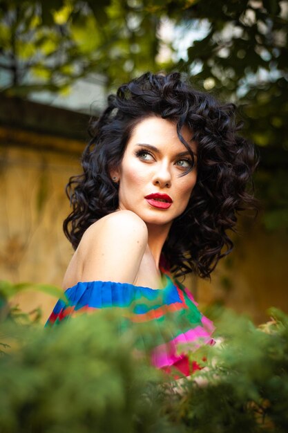 A woman with curly hair stands in front of a wall with trees in the background.