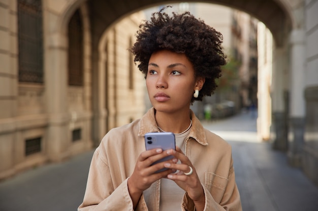 Donna con i capelli ricci usa il cellulare legge contenuti dai social network indossa giacca beige posa fuori naviga internet manda sms