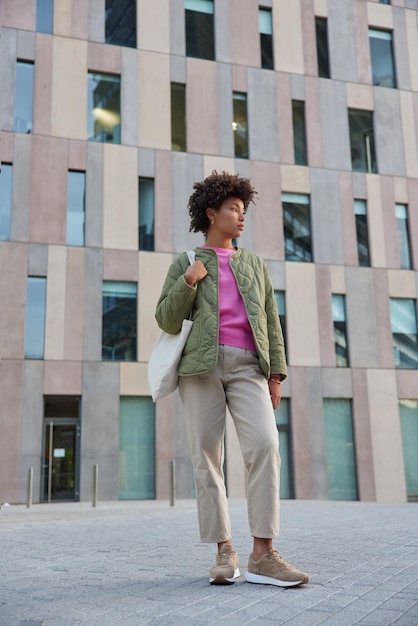Donna con i capelli ricci indossa giacca giacca pantaloni e scarpe da ginnastica porta borsa in tessuto pone all'aperto contro il moderno edificio urbano cammina fuori