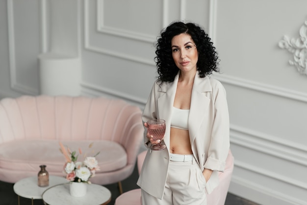 Photo woman with dark curly hair in a light suit with a naked belly holds a glass in her hands